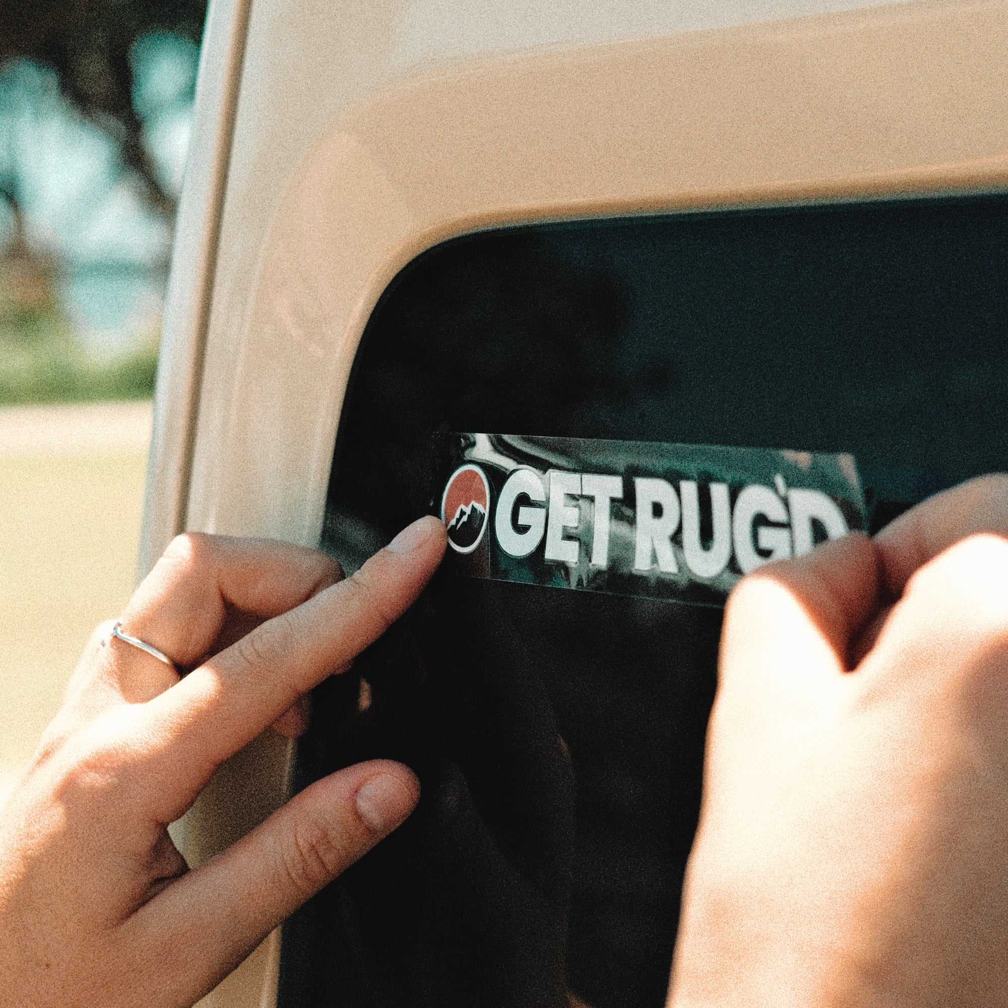 Hands applying a Get Rug'd car decal sticker from the coaster pack to a vehicle window
