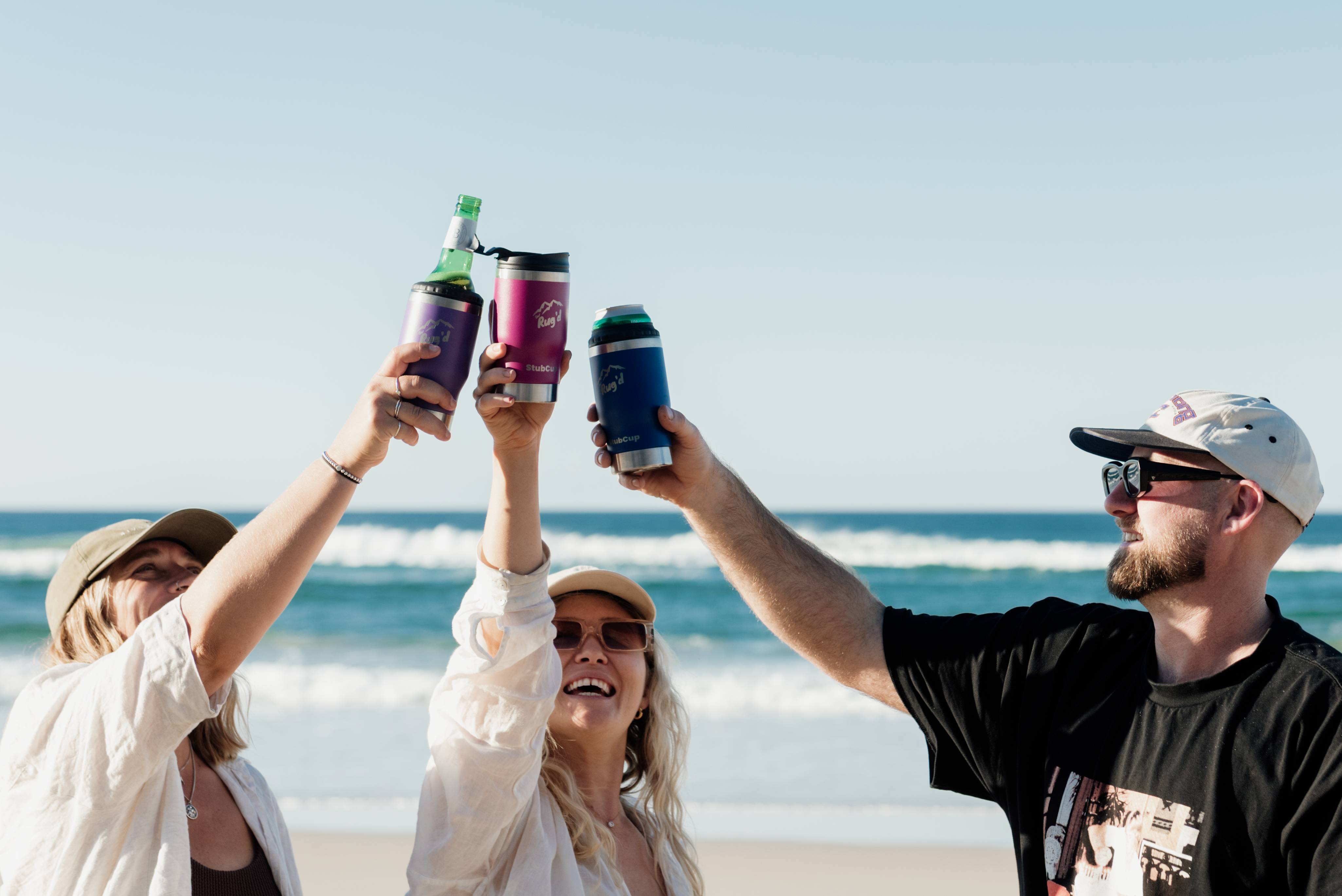 Three people on a beach holding drinks in cans, with a clear blue sky and ocean in the background.