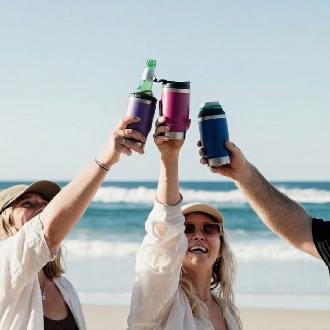 Three friends toasting with RUGD StubCup insulated stubby coolers on an Australian beach — purple, pink and navy colours holding beer bottles and cans