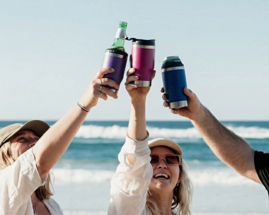 Three friends toasting with RUGD StubCup insulated stubby coolers on an Australian beach — purple, pink and navy colours holding beer bottles and cans