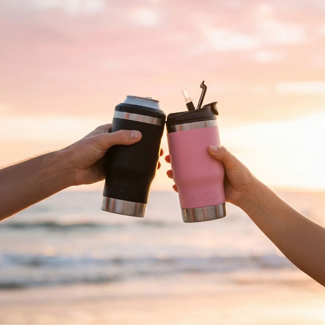 Two RUGD StubCups toasting at sunset on an Australian beach — black stubby cooler holding a can and pink insulated tumbler with straw lid