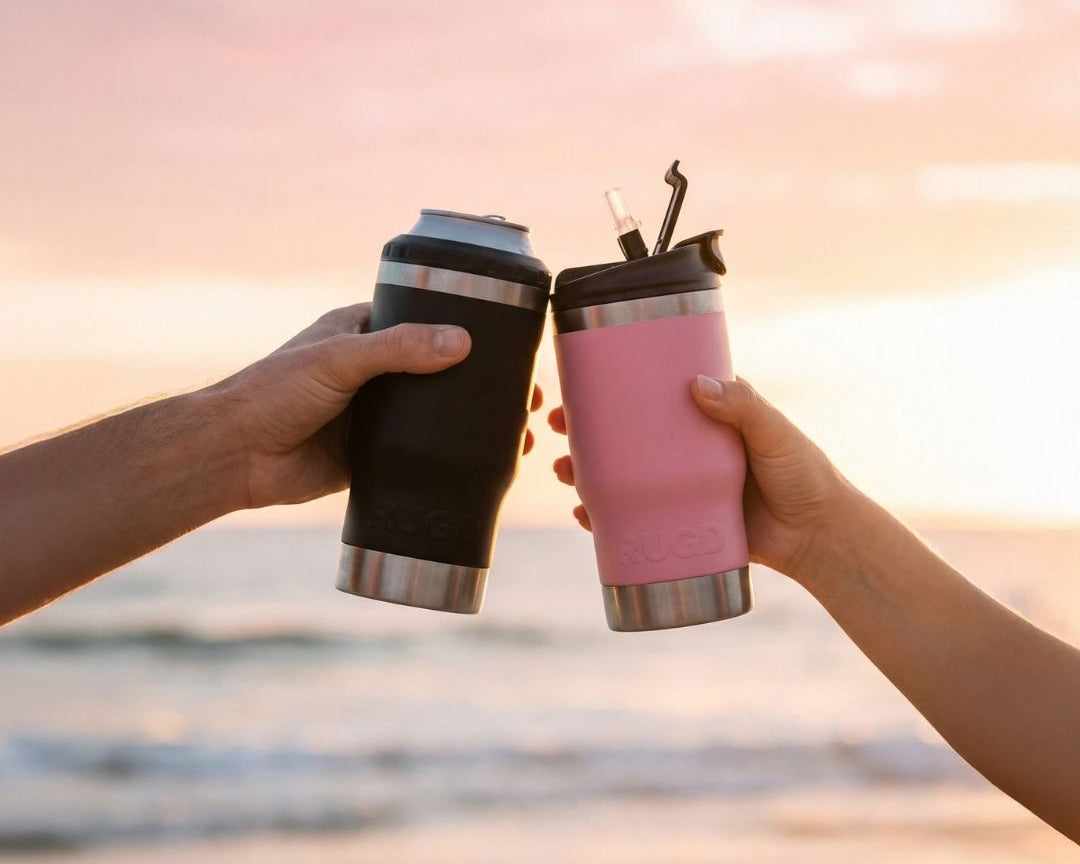Two RUGD StubCups toasting at sunset on an Australian beach — black stubby cooler holding a can and pink insulated tumbler with straw lid