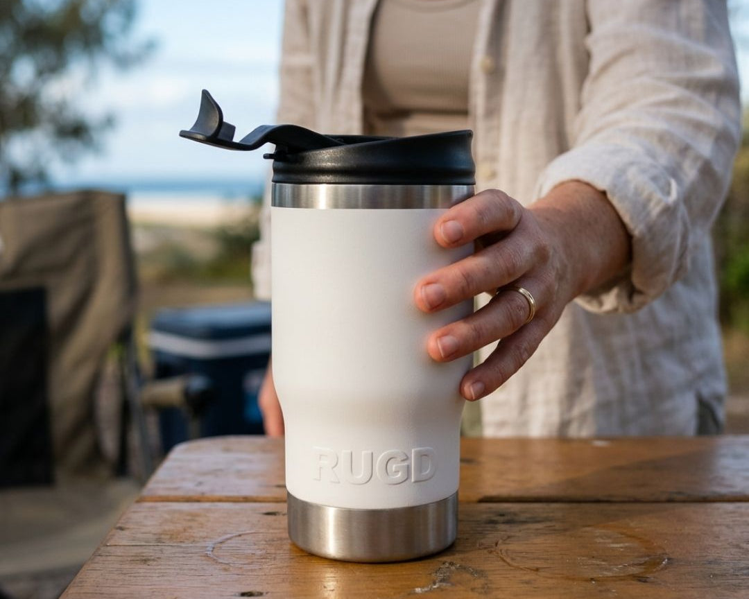 Woman picking up a white RUGD StubCup with SwigSip coffee lid from a wooden outdoor table at a beachside campsite