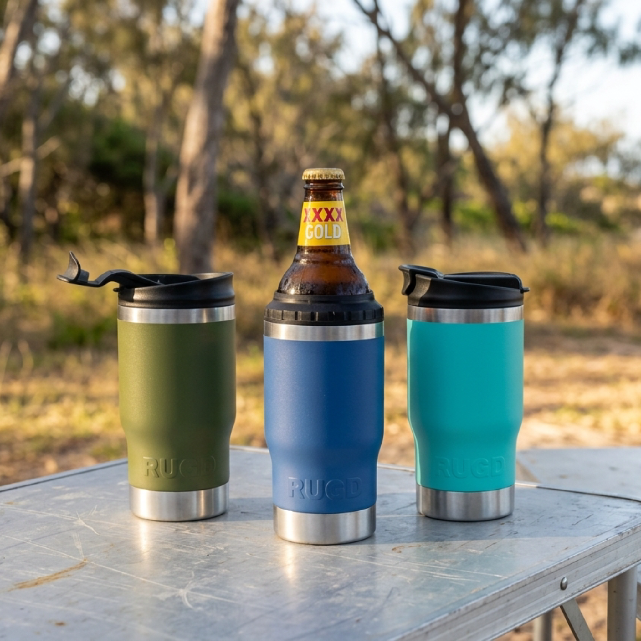 Three insulated travel mugs in green, blue, and teal with a bottle on a table outdoors.