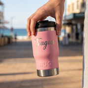 Person holding a pink insulated tumbler with 'Teagan' and 'RUGD' branding outdoors.