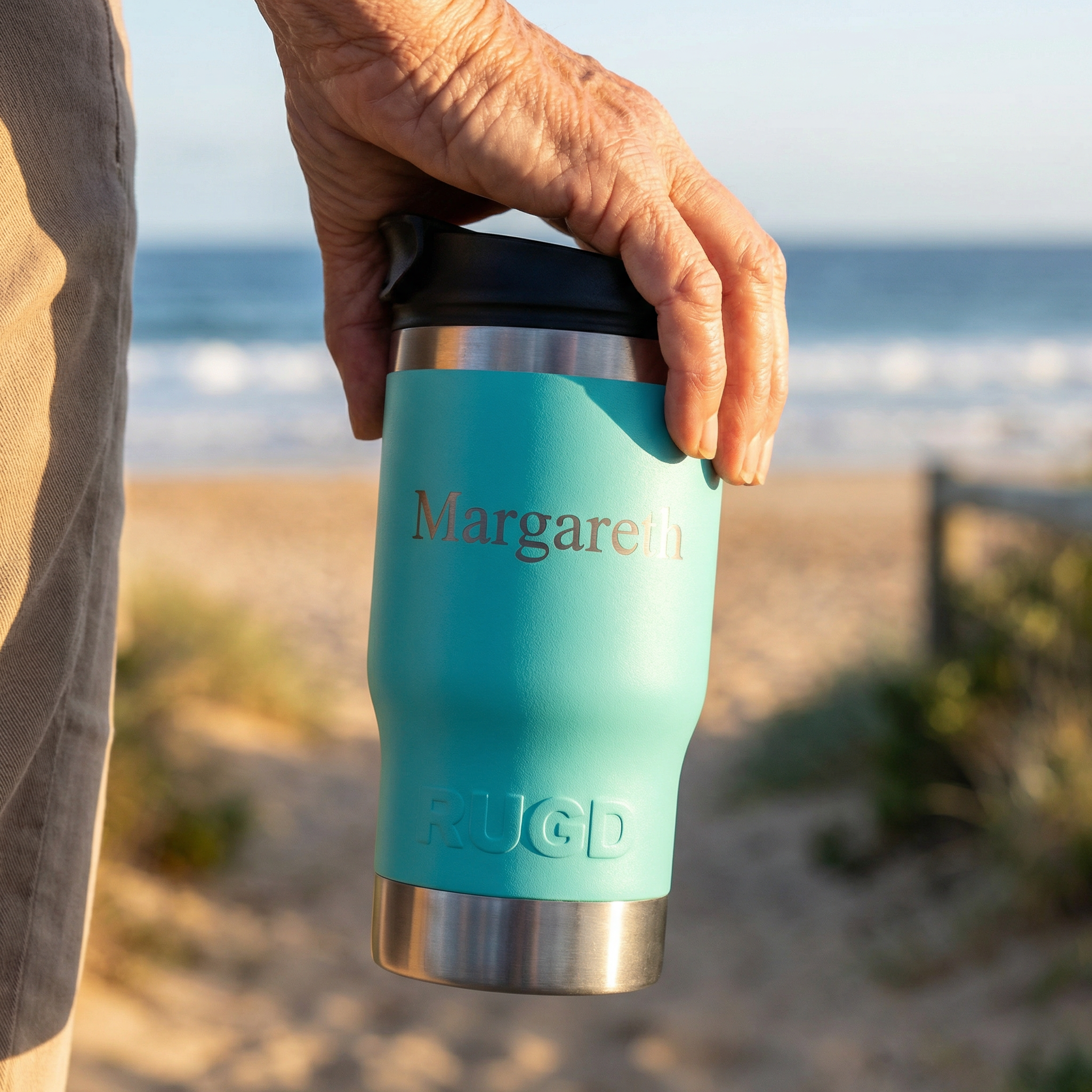 Person holding a teal mug with 'Margaretha' on it at the beach