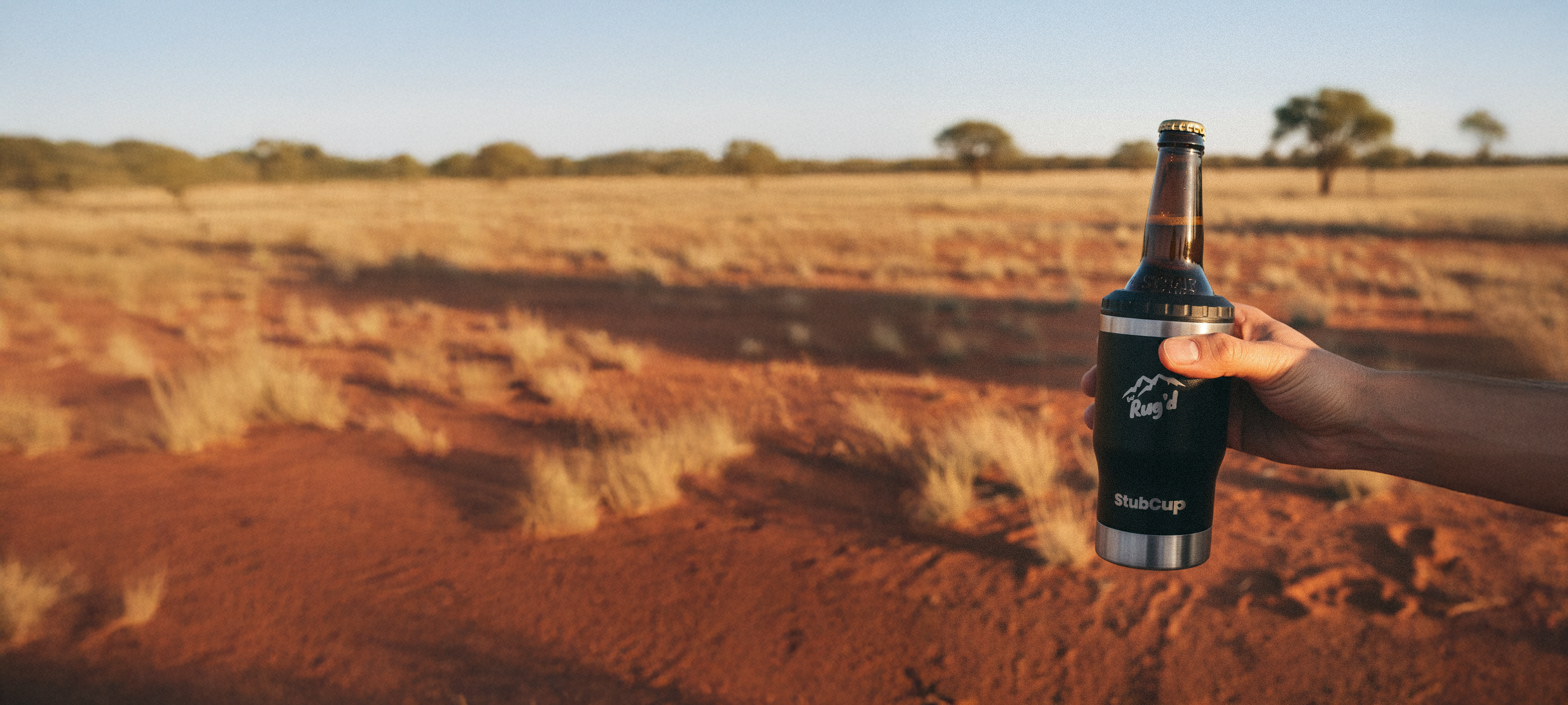 Person holding a beer bottle with a tumbler in a desert landscape