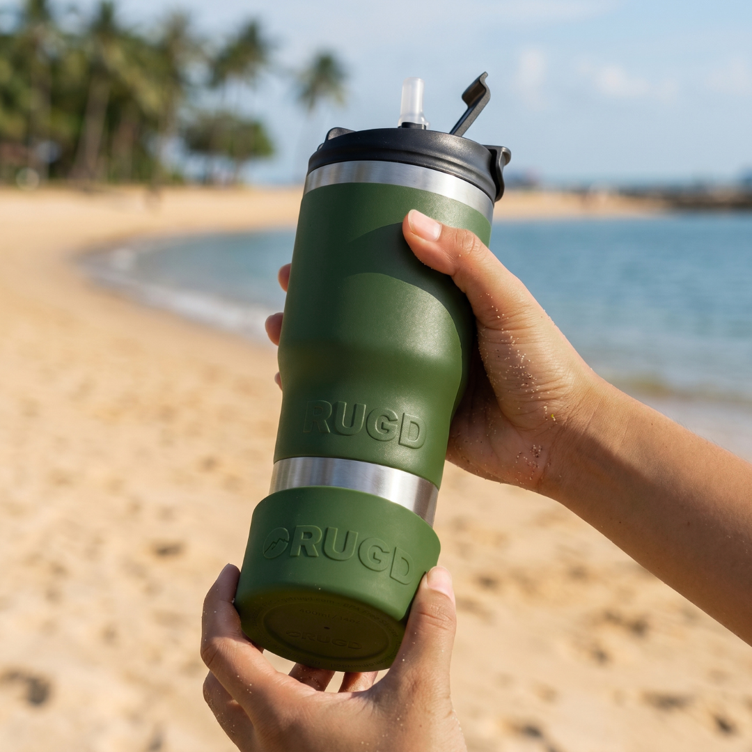 A person holds a green insulated tumbler with a black lid and clear straw at a beach, with sand, water, and palm trees blurred in the background. The tumbler has embossed 'RUGD' branding.