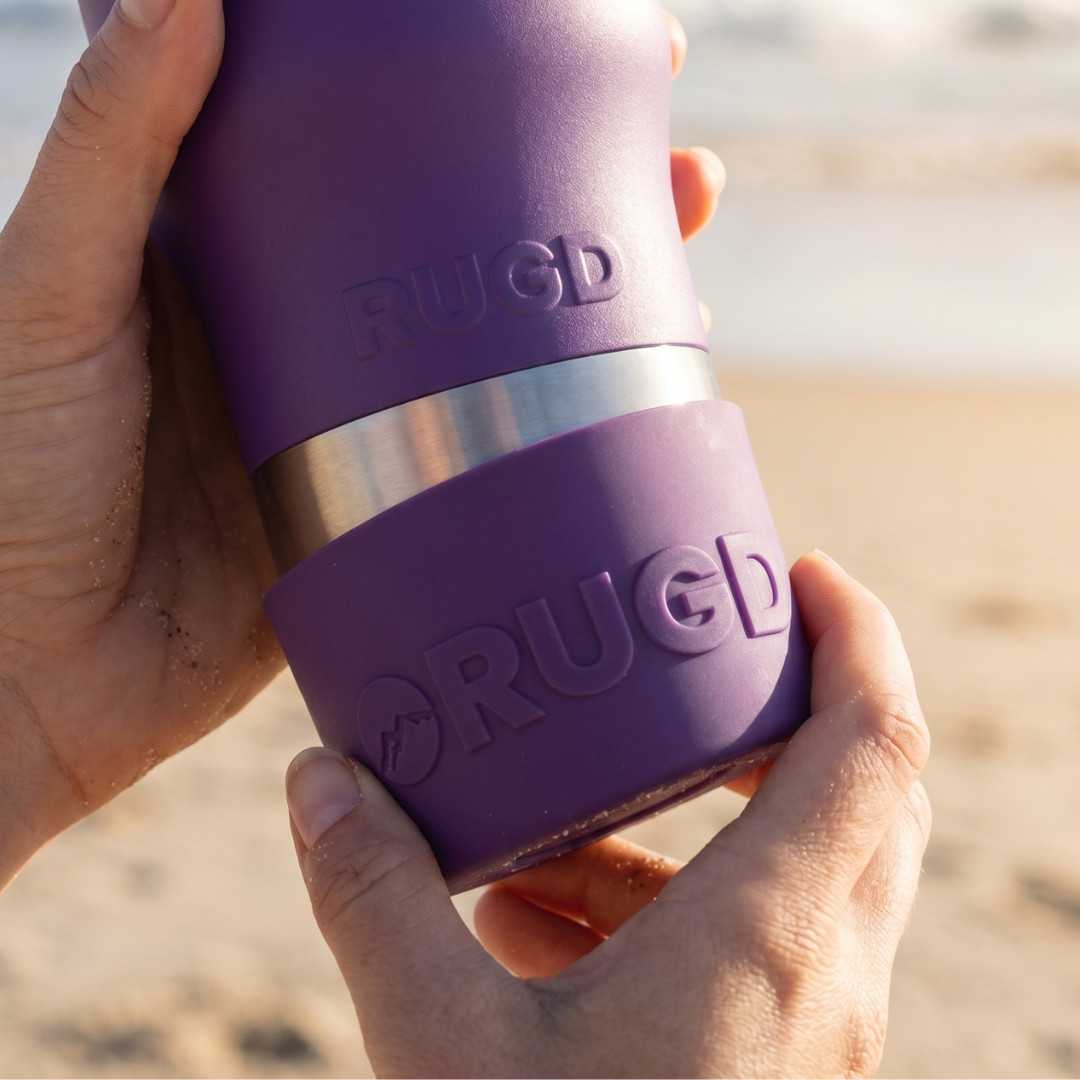 A close-up of a person holding a purple RUGD insulated tumbler with a stainless steel band, set against a blurred beach background.