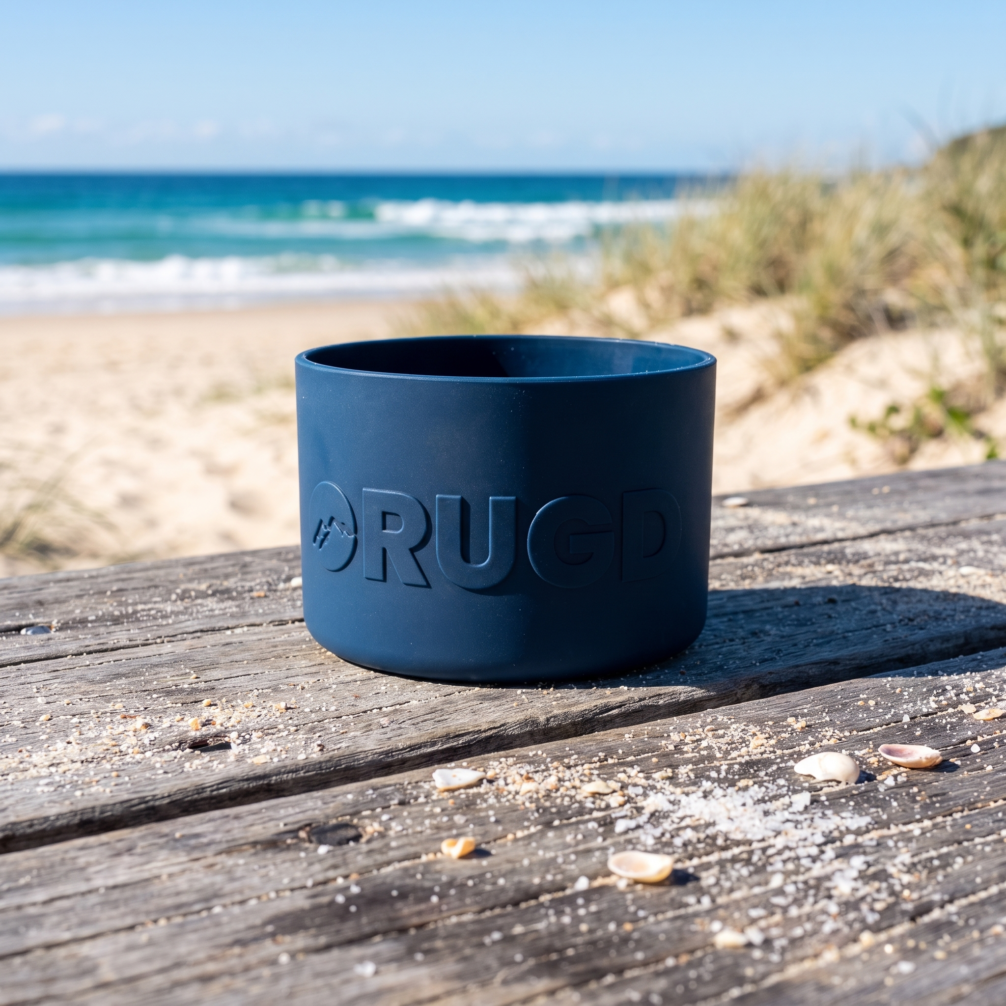 A dark blue cylindrical silicone boot holder with the brand name 'RUGD' embossed on the front, sitting on a weathered wooden table with a beach and ocean in the blurred background.