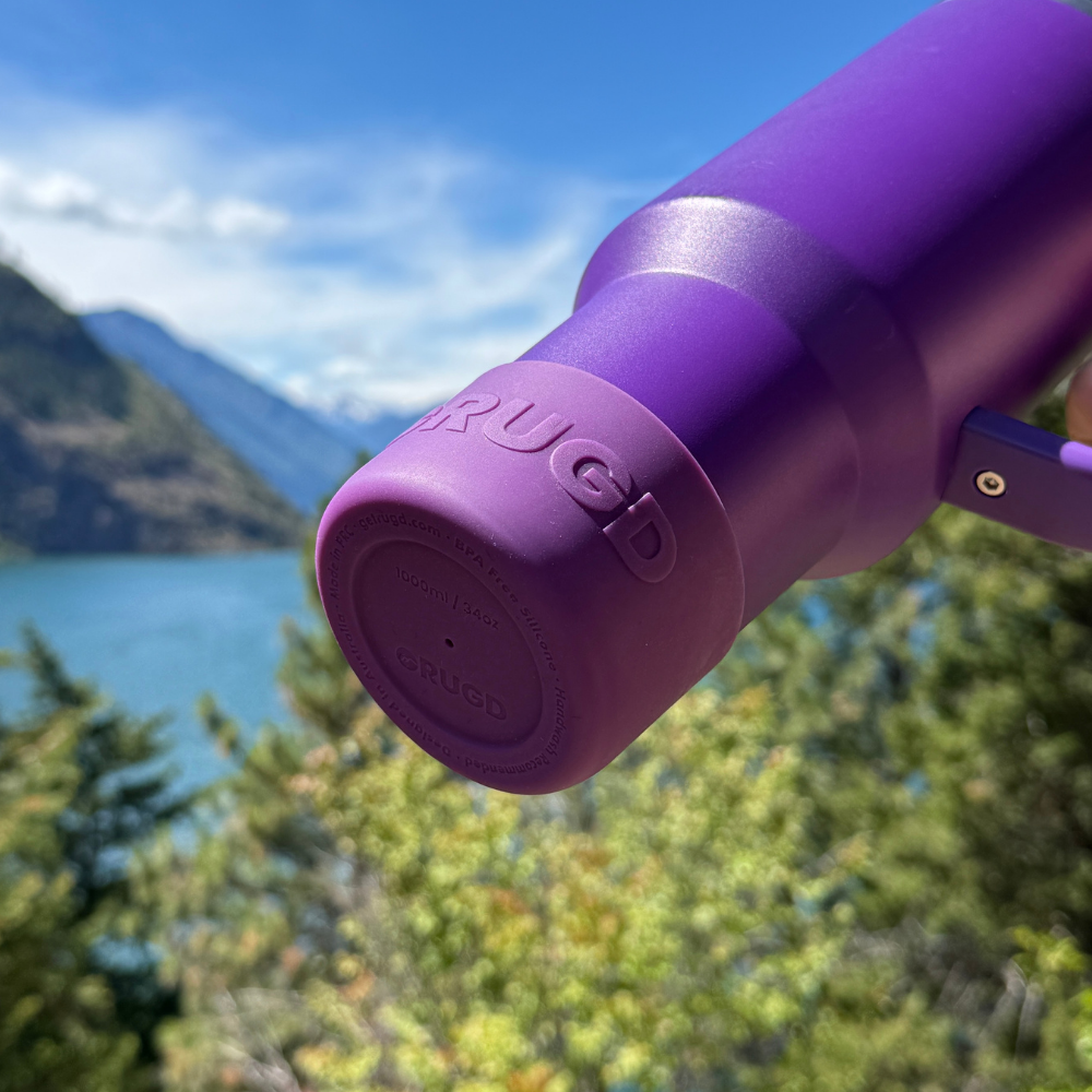 A close-up of a purple insulated bottle or tumbler with the brand name 'CRUIC' embossed on the cap, set against a blurred outdoor background of a lake, mountains, and trees.