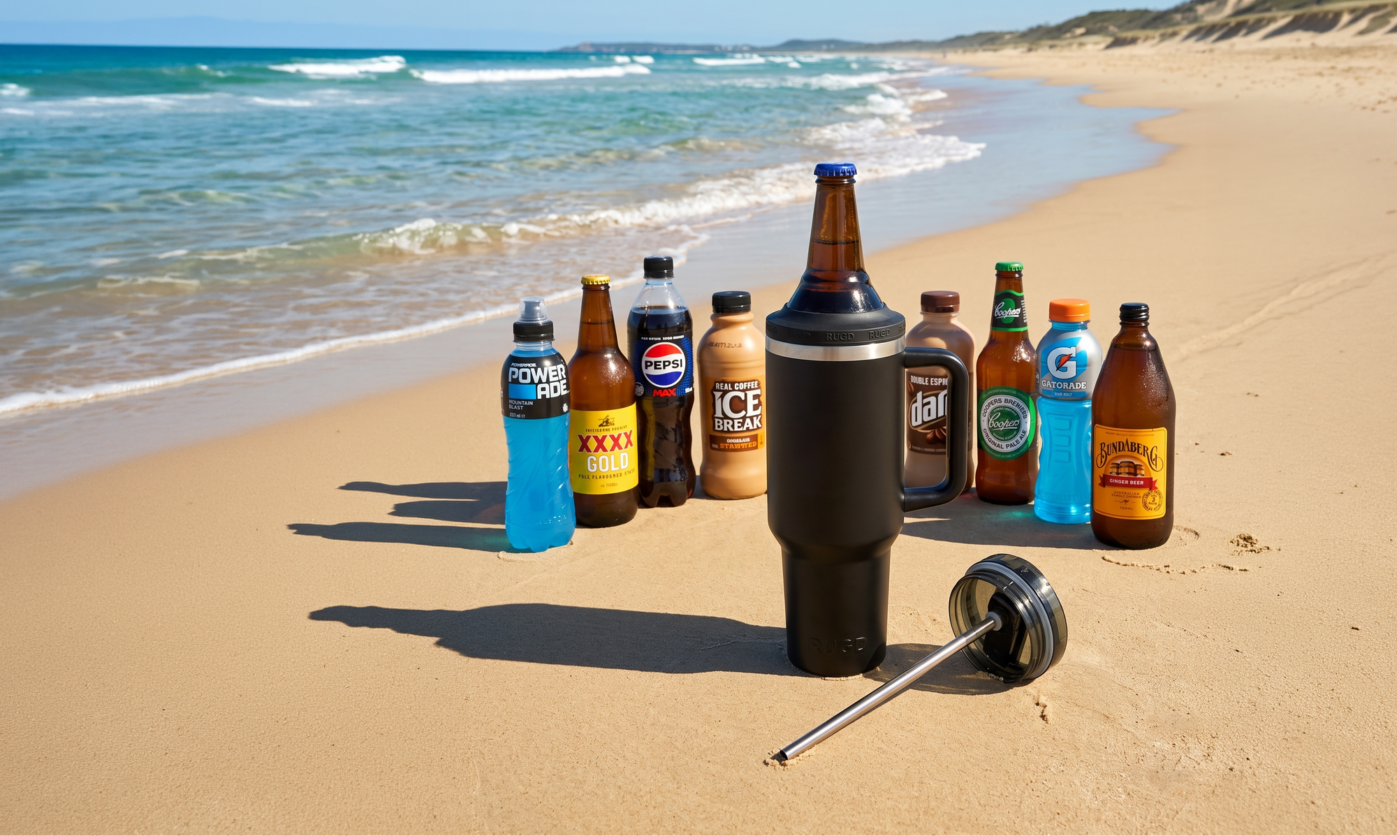 A group of beverage bottles and cans arranged on a sandy beach next to the ocean, with a metal strainer resting in the sand in the foreground. The scene includes visible ocean waves, blue sky, and dunes in the background.